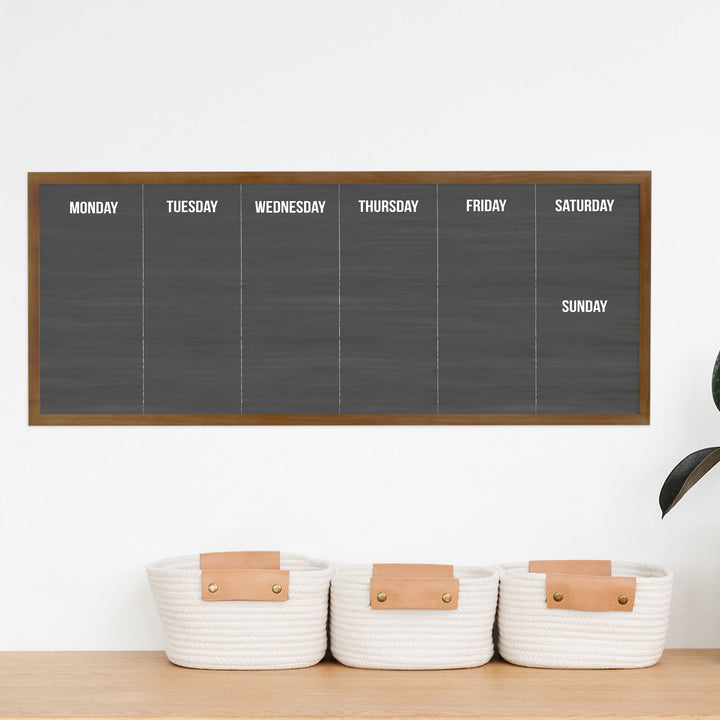 A framed slim dry-erase weekly calender with a faux chalkboard look hanging on the wall