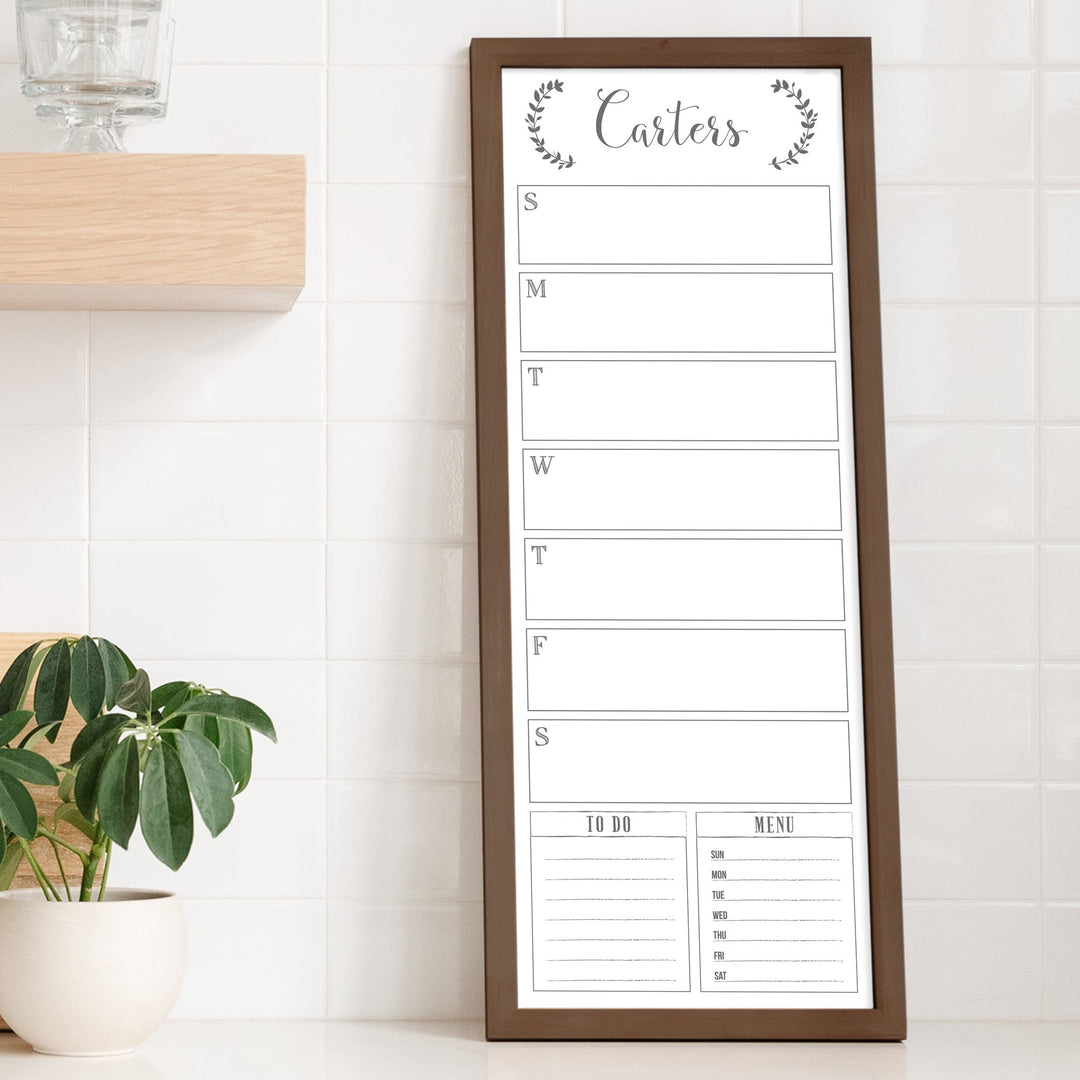 A framed slim dry-erase weekly calender with a faux chalkboard look hanging on the wall