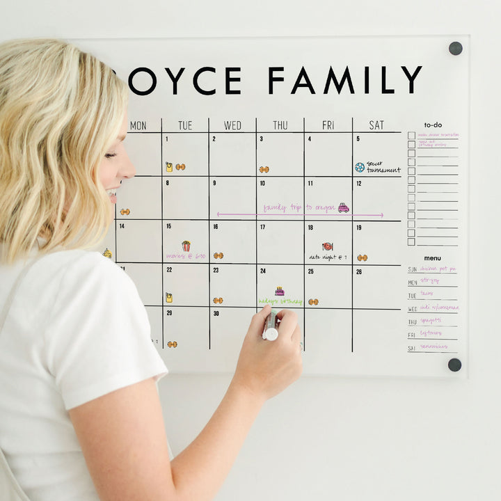 a woman uses brightly colored chalk markers and resuable schedule stickers on an acrylic calendar