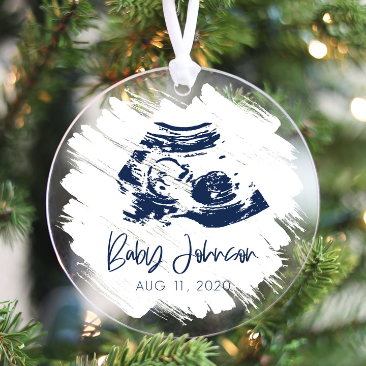A Christmas ornament with a navy colored ultrasound on a white brushed background.