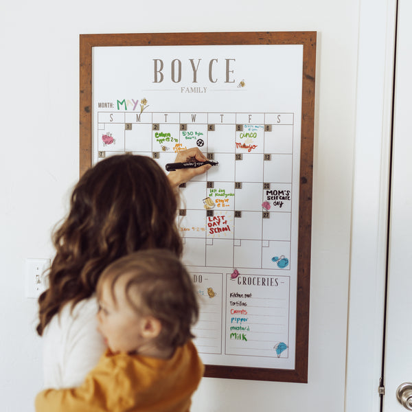 Woman holding baby writing on a dry-erase monthly calendar. The calendar is framed and personalized with a family name and is also magnetic with magnetic date numbers.