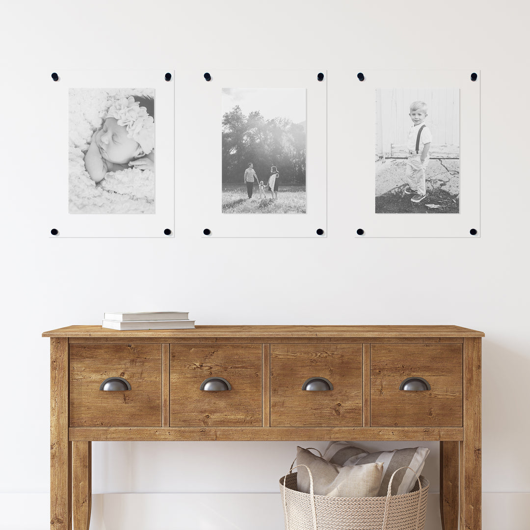a white wall displays three floating acrylic picture frames with black hardware. They are hanging over a classic wooden entryway table. 