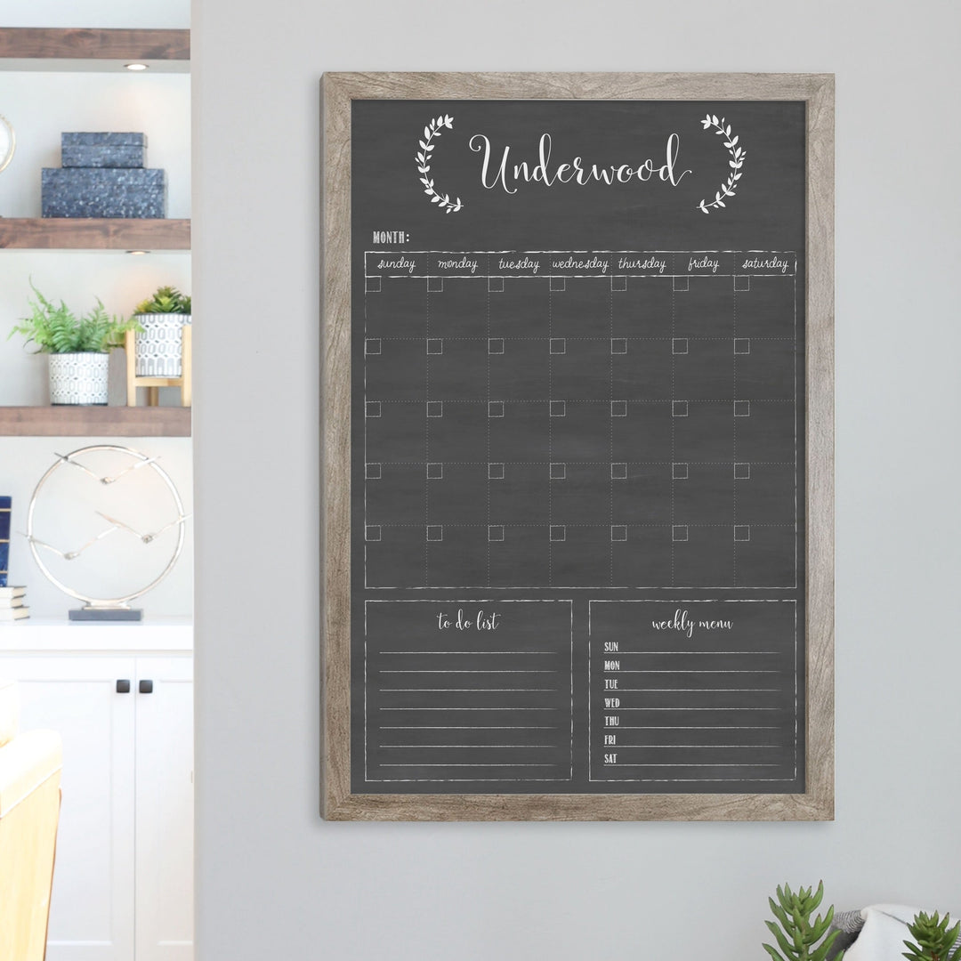 A framed dry-erase monthly calender with a chalkboard look hanging on the wall