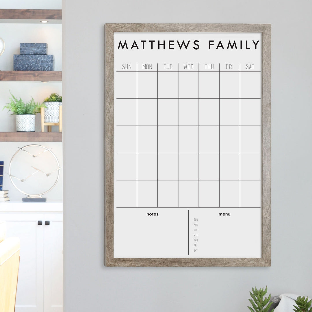 A framed dry-erase monthly calender with a chalkboard look hanging on the wall