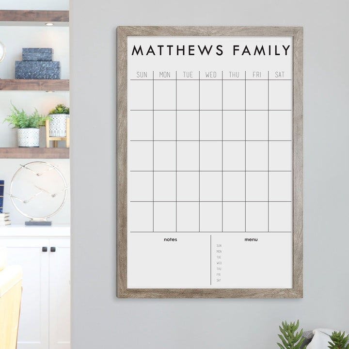 A framed dry-erase monthly calender with a chalkboard look hanging on the wall