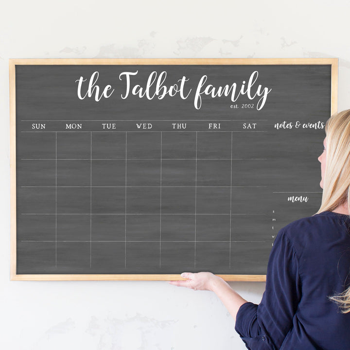 A framed dry-erase monthly calender with a chalkboard look hanging on the wall
