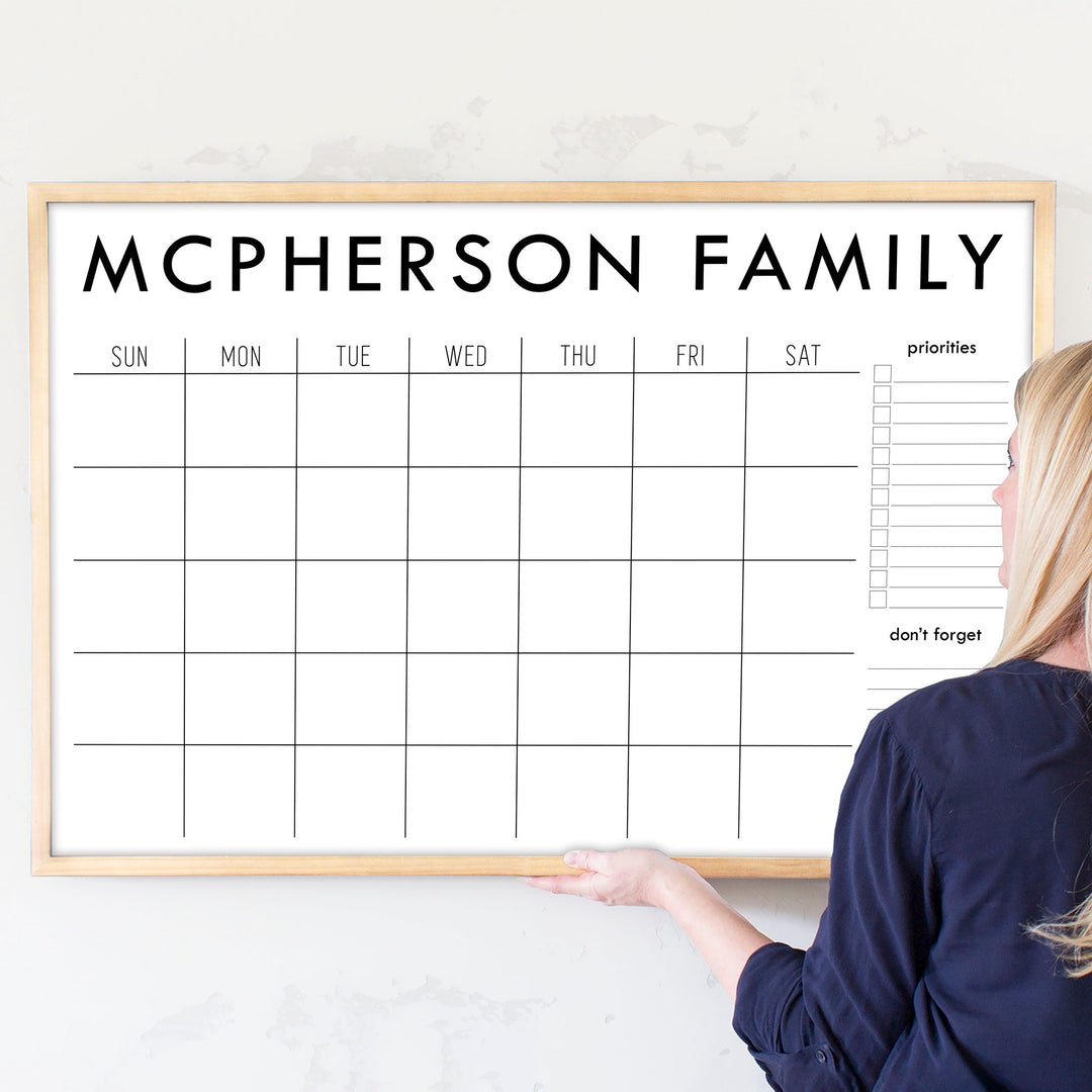 A framed dry-erase monthly calender with a chalkboard look hanging on the wall