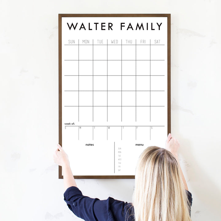 A framed whiteboard calendar with a monthly and weekly format hanging on the wall