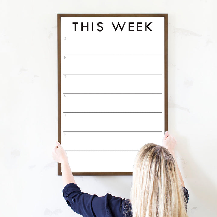 A framed whiteboard weekly calender hanging on the wall