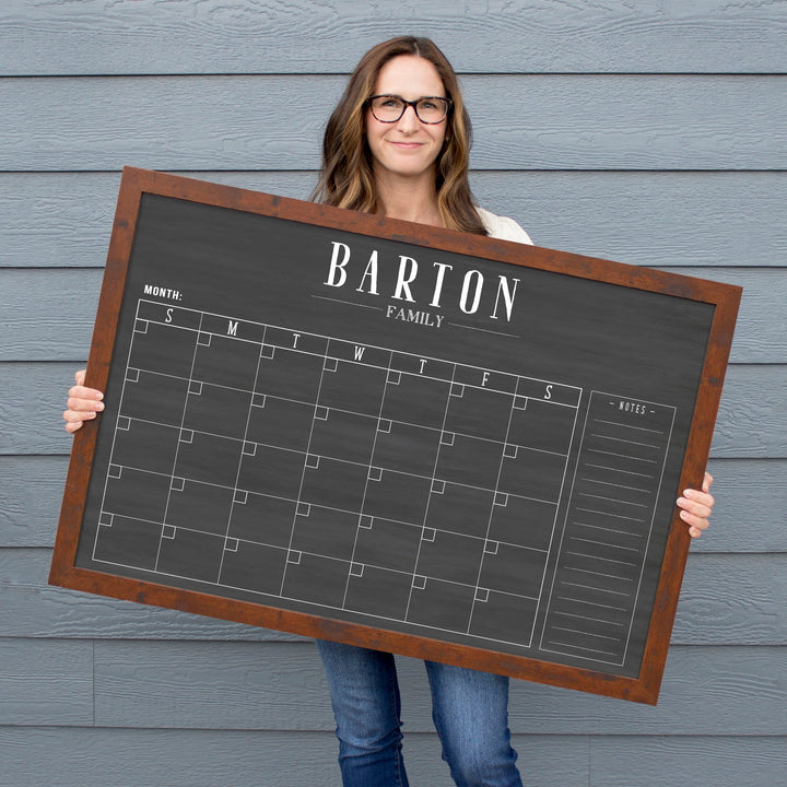 A framed dry-erase monthly calender with a chalkboard look hanging on the wall