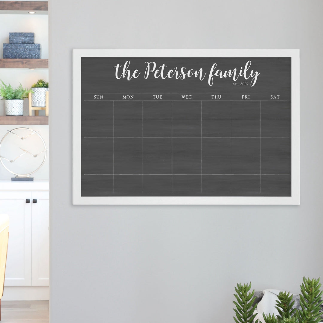 A framed dry-erase monthly calender with a chalkboard look hanging on the wall