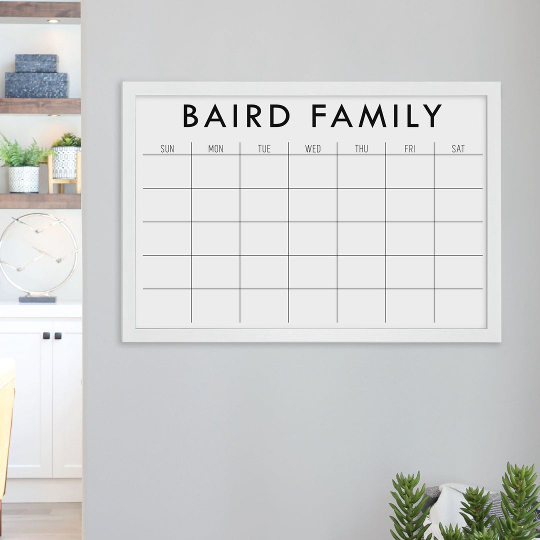 A framed dry-erase monthly calender with a chalkboard look hanging on the wall