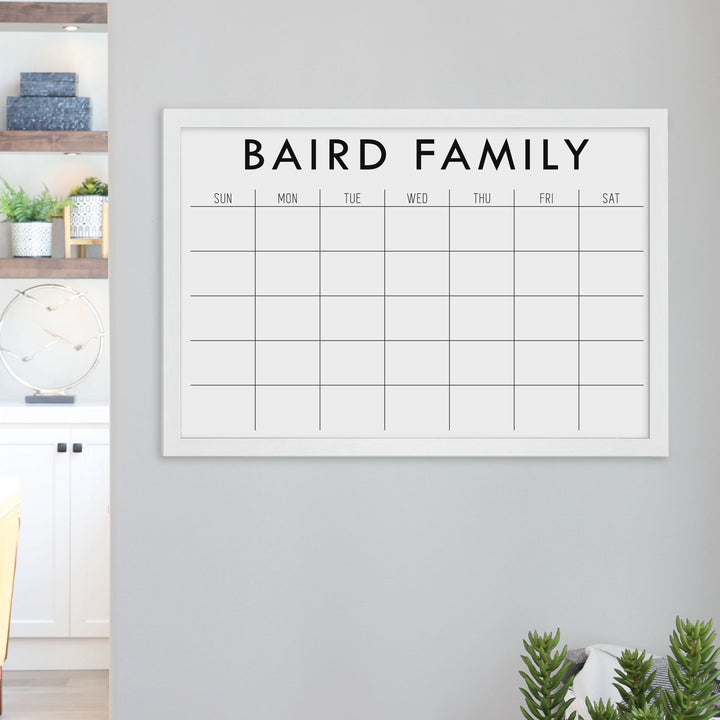 A framed dry-erase monthly calender with a chalkboard look hanging on the wall