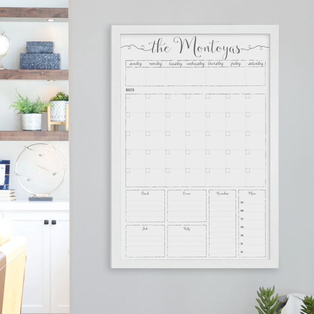 A framed whiteboard calendar with a monthly and weekly format hanging on the wall