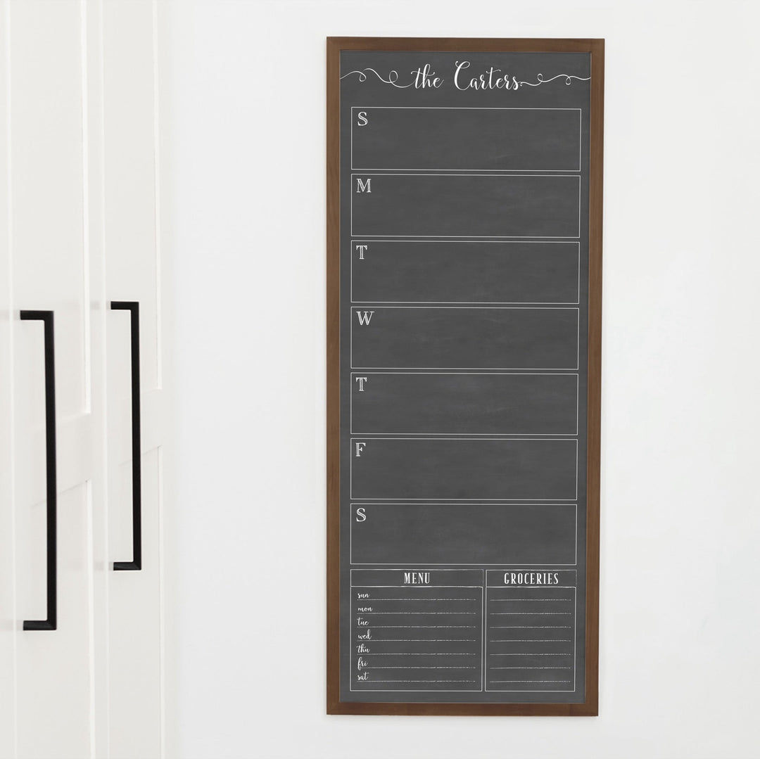 A framed slim dry-erase weekly calender with a faux chalkboard look hanging on the wall