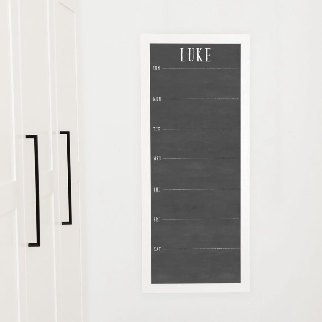 A framed slim dry-erase weekly calender with a faux chalkboard look hanging on the wall