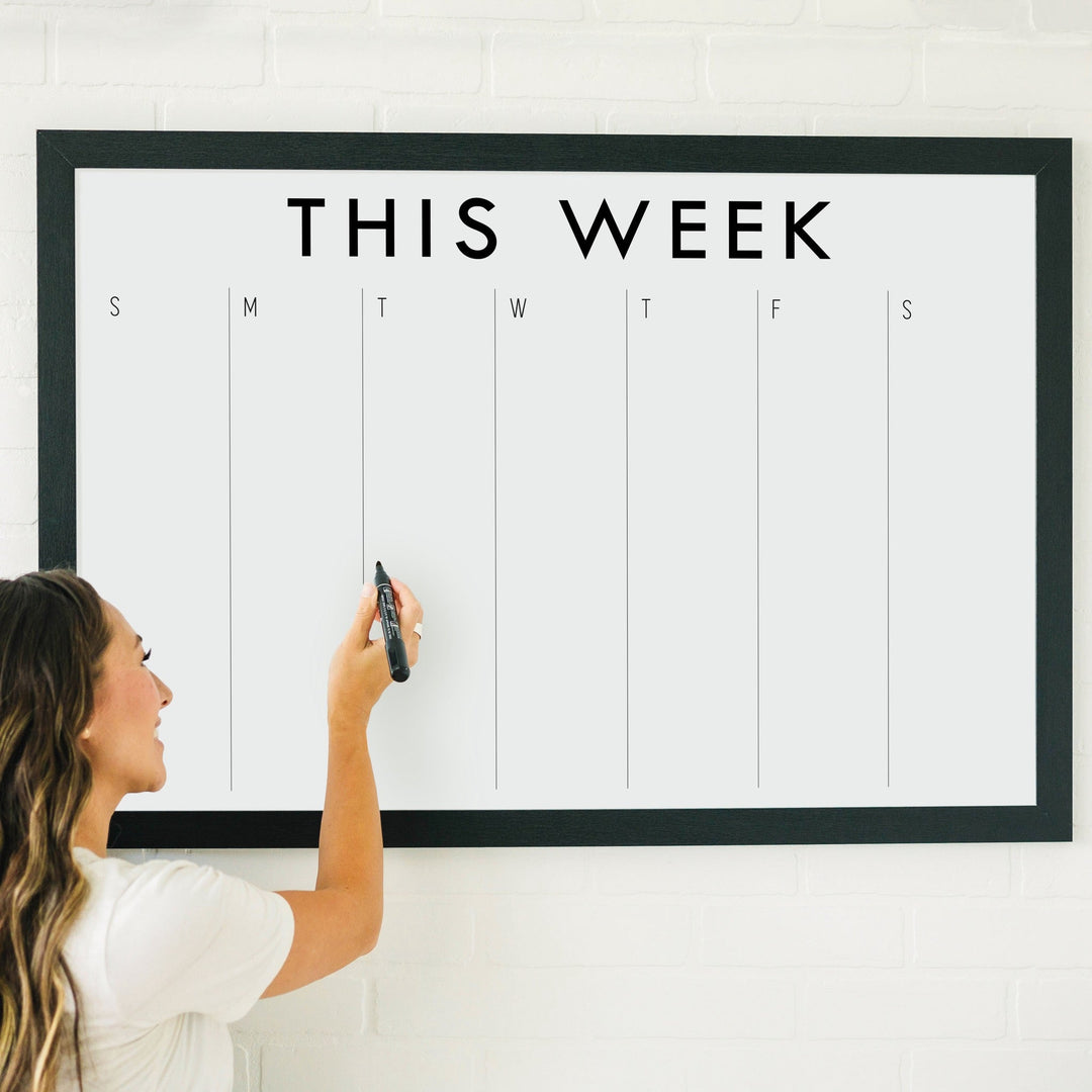 A framed whiteboard weekly calender hanging on the wall