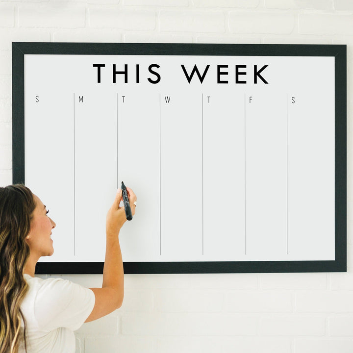 A framed whiteboard weekly calender hanging on the wall