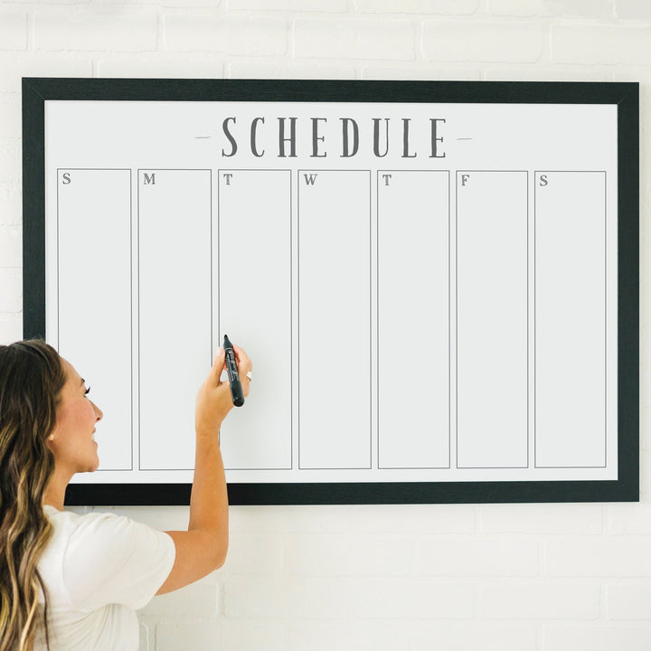 A framed whiteboard weekly calender hanging on the wall