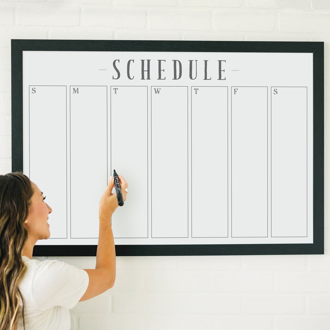 A framed whiteboard weekly calender hanging on the wall