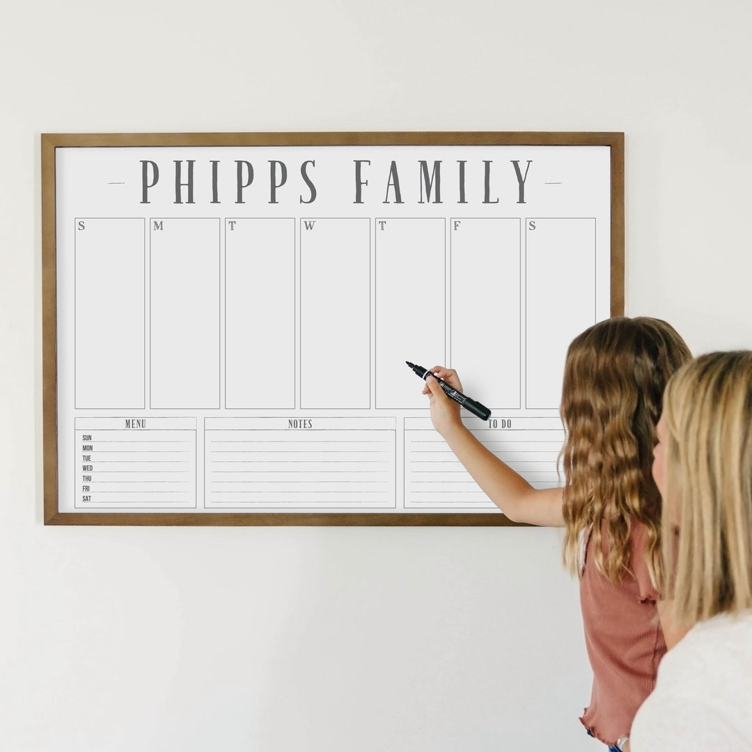 A framed whiteboard weekly calender hanging on the wall