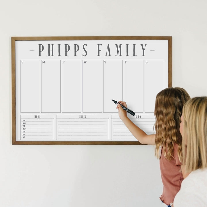 A framed whiteboard weekly calender hanging on the wall