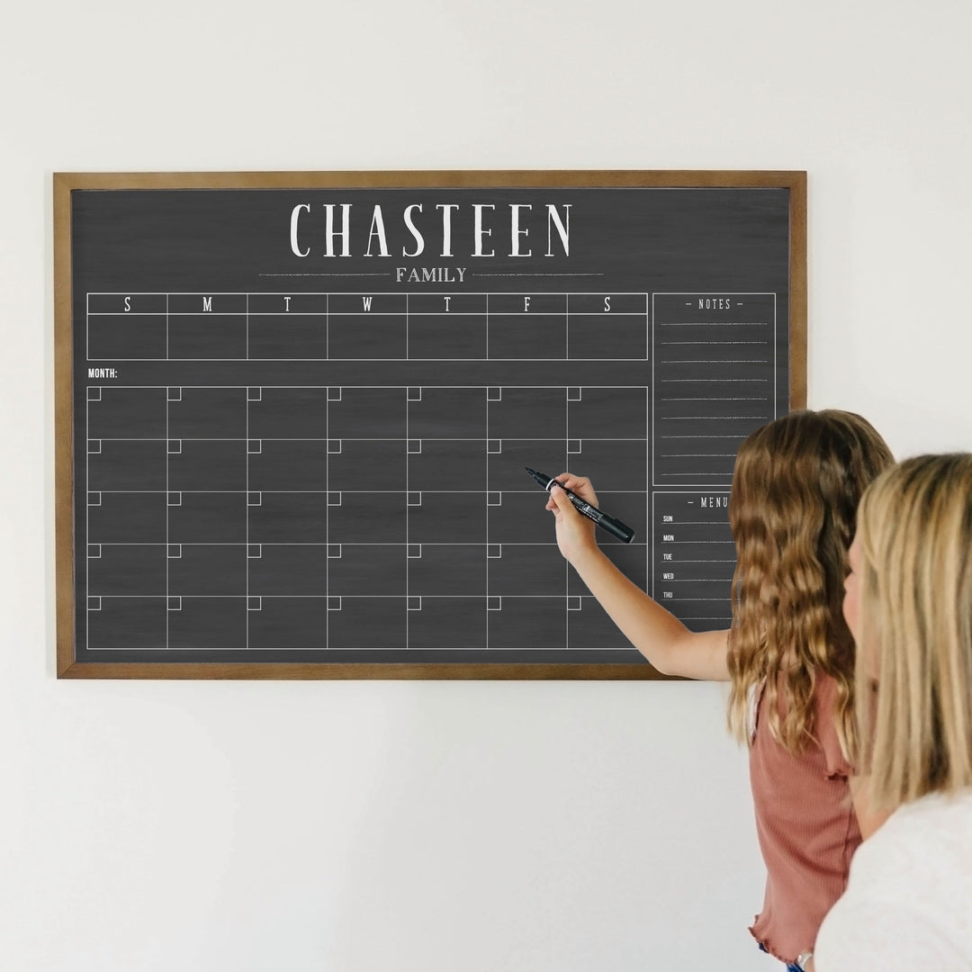 A framed dry-erase chalkboard calendar with a monthly and weekly format hanging on the wall