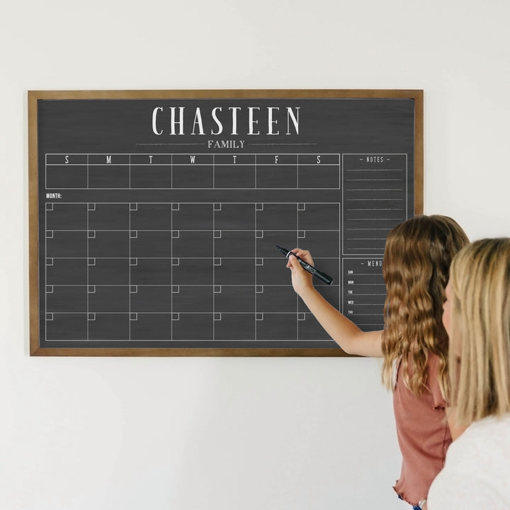 A framed dry-erase chalkboard calendar with a monthly and weekly format hanging on the wall