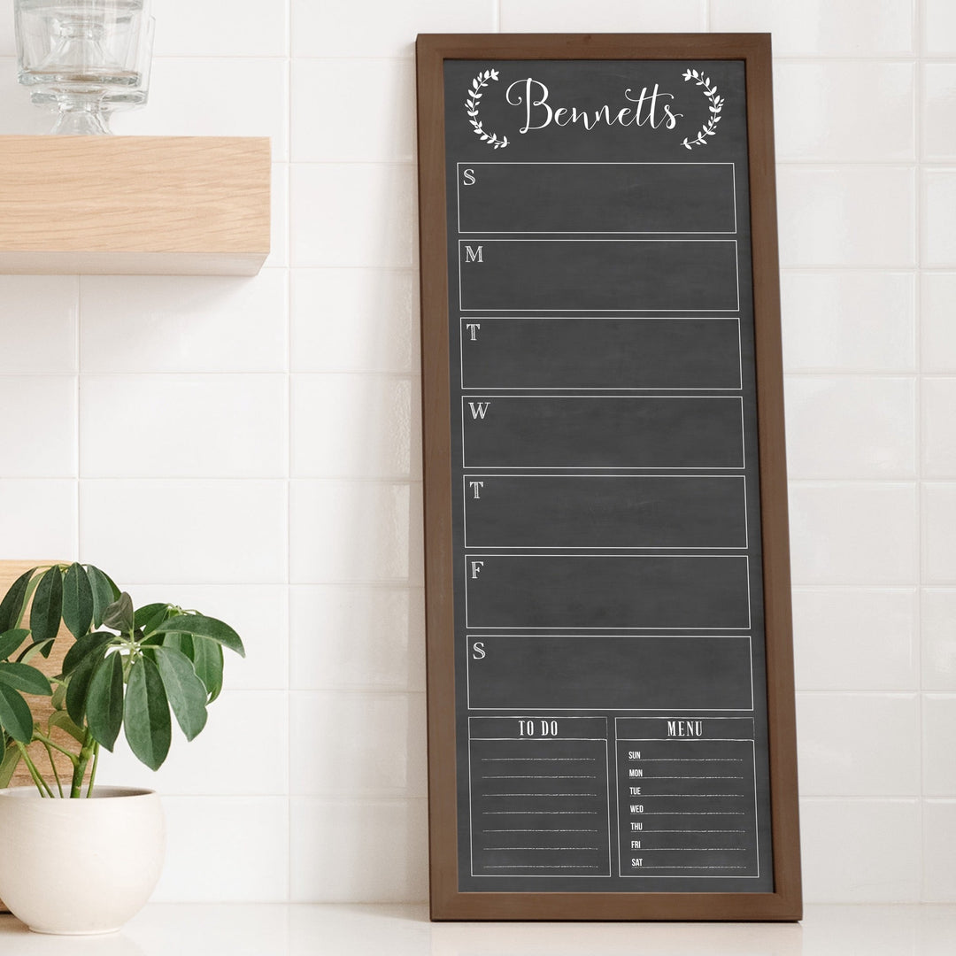 A framed slim dry-erase weekly calender with a faux chalkboard look hanging on the wall