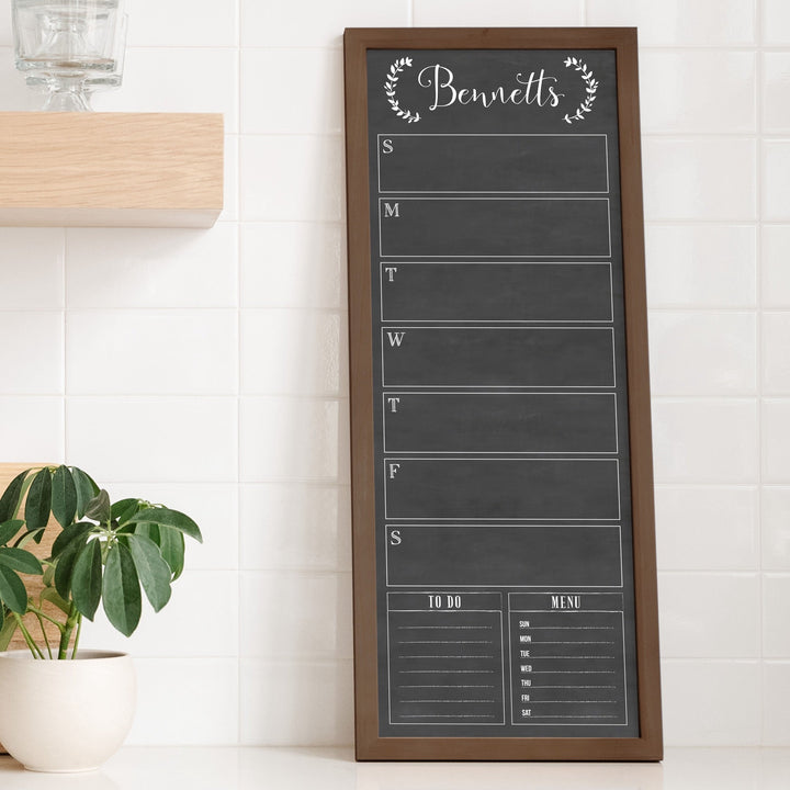 A framed slim dry-erase weekly calender with a faux chalkboard look hanging on the wall