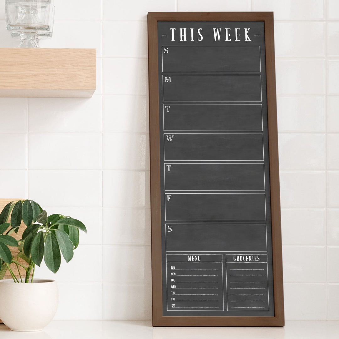 A framed slim dry-erase weekly calender with a faux chalkboard look hanging on the wall