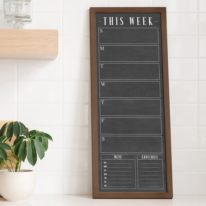 A framed slim dry-erase weekly calender with a faux chalkboard look hanging on the wall