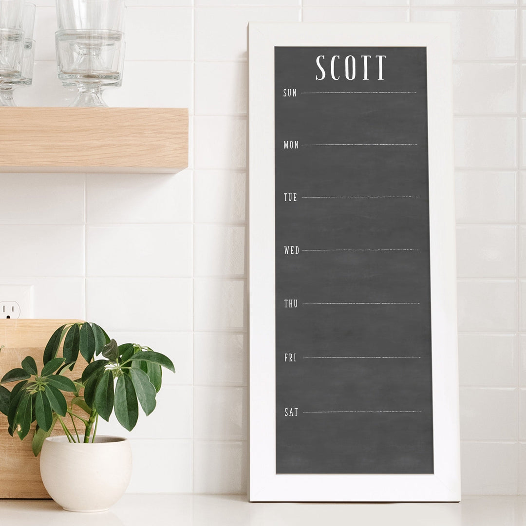 A framed slim dry-erase weekly calender with a faux chalkboard look hanging on the wall
