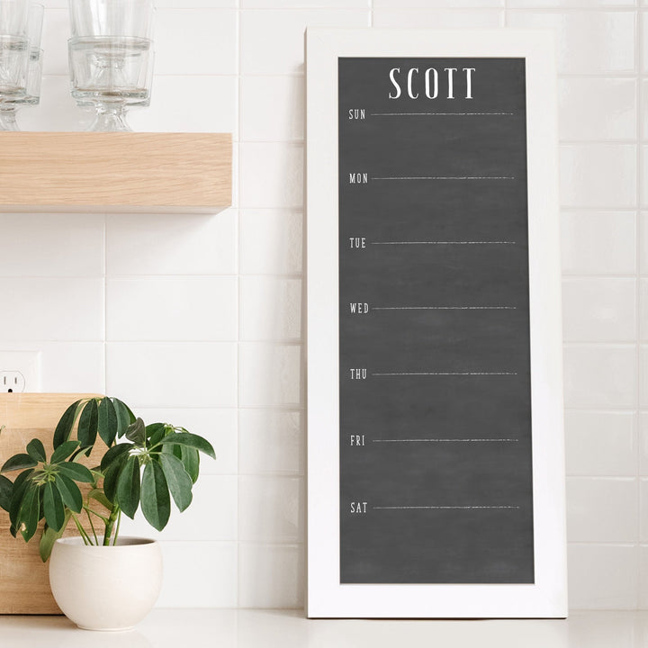 A framed slim dry-erase weekly calender with a faux chalkboard look hanging on the wall
