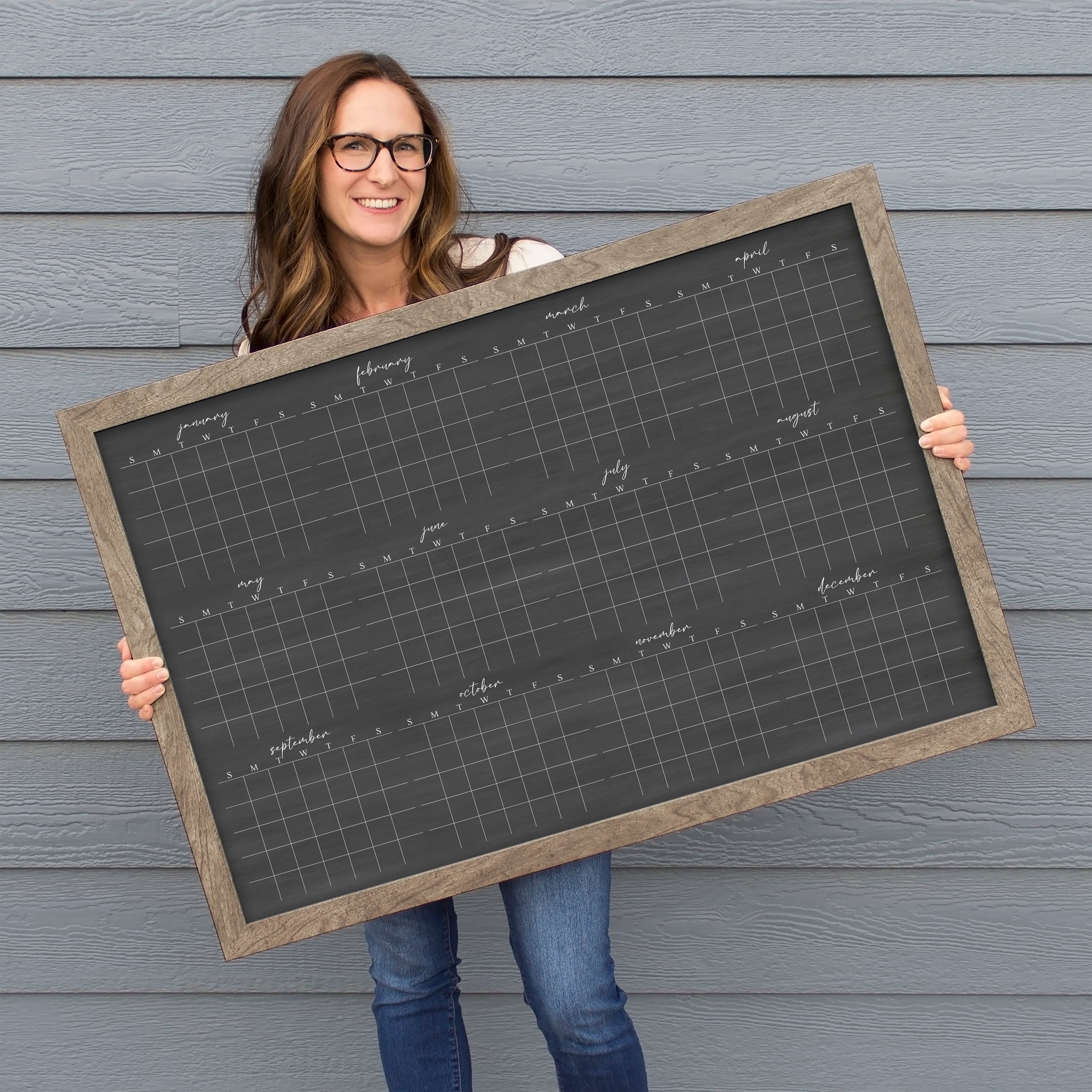 A framed dry-erase yearly calendar with a chalkboard look hanging up on the wall