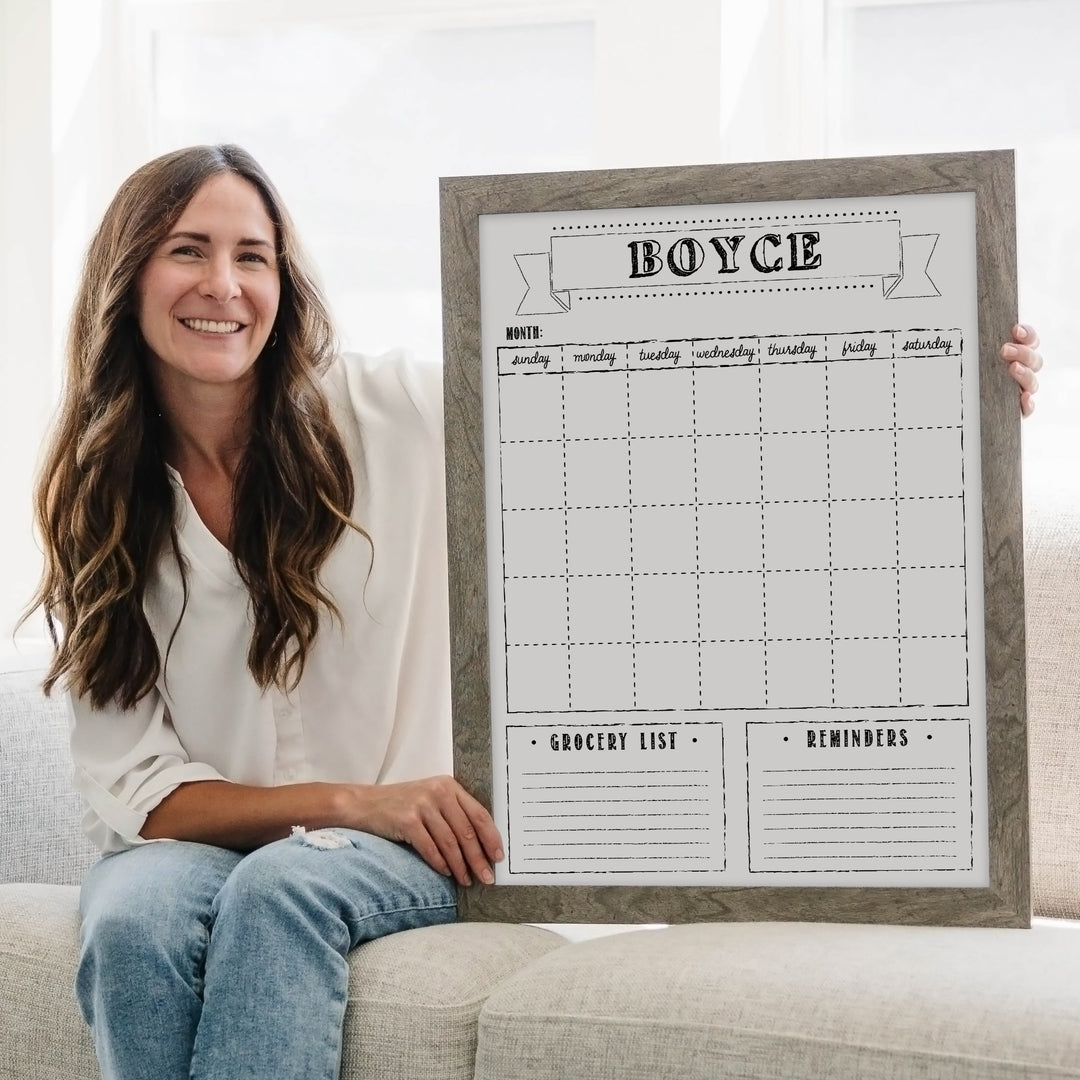 Woman holding a large framed calendar on a couch in a bright room.