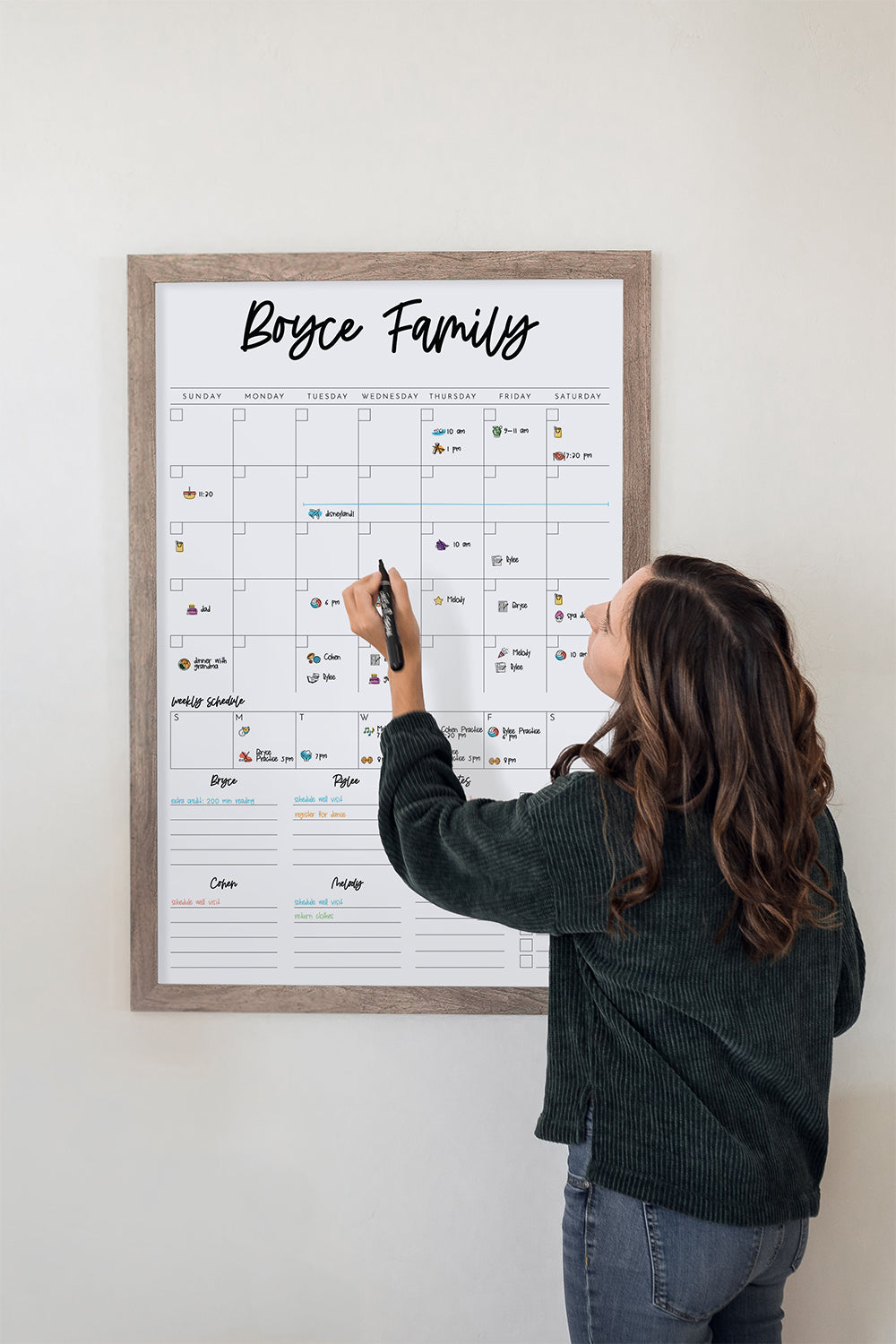 Woman writing on a framed dry-erase family calendar titled 'Boyce Family' against a white wall. The calendar has sticker icons on it to represent different activities.
