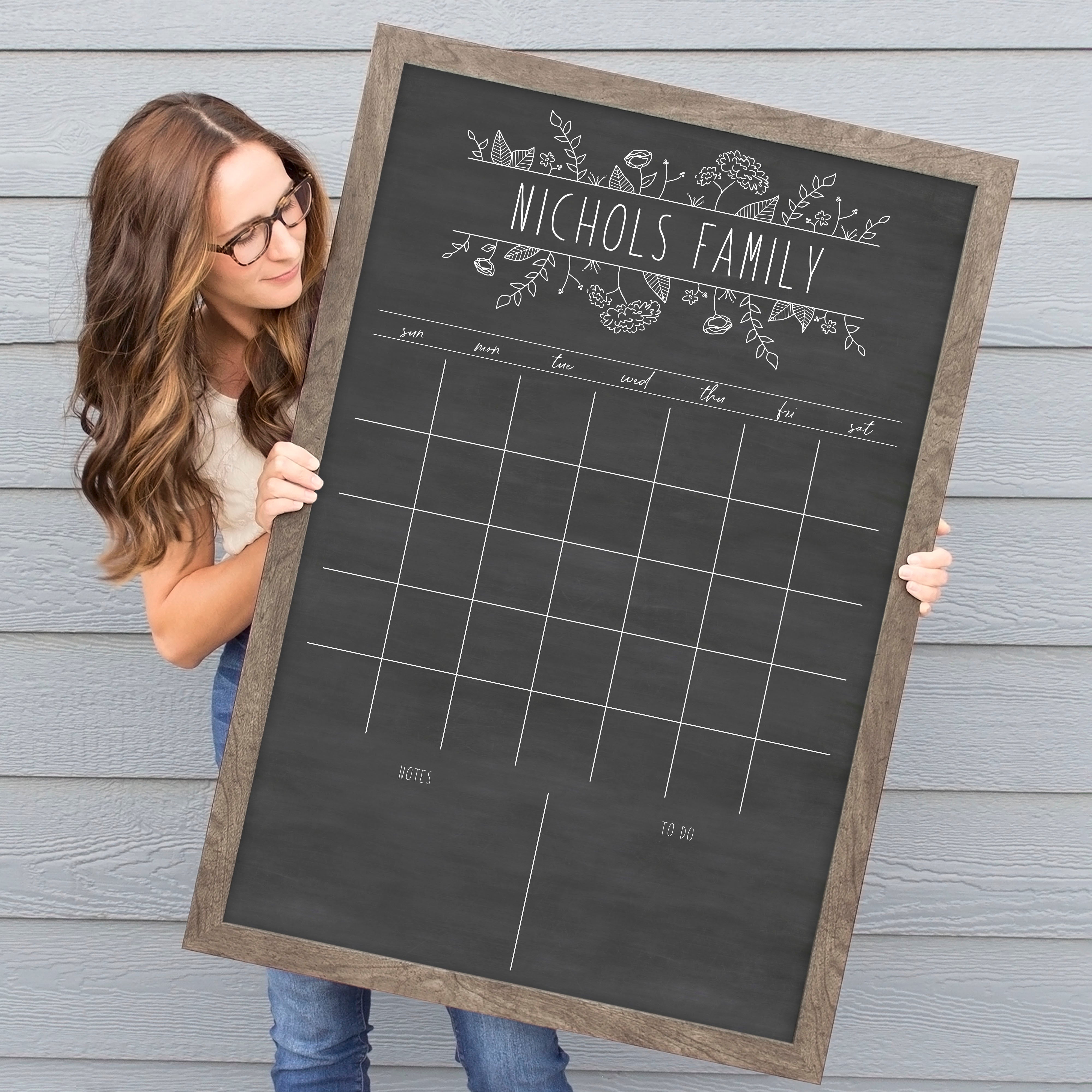 A framed dry-erase monthly calender with a chalkboard look hanging on the wall