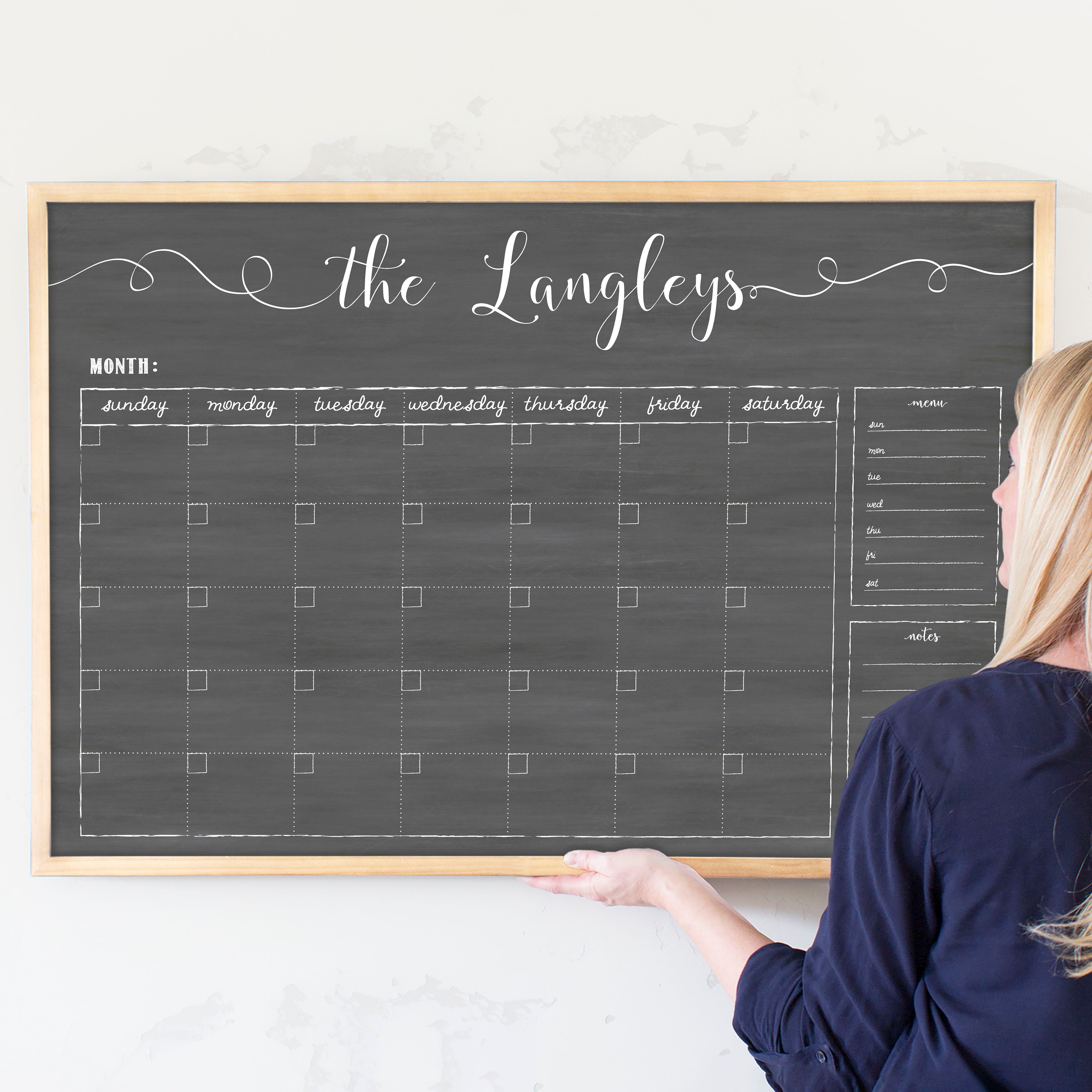 A framed dry-erase monthly calender with a chalkboard look hanging on the wall