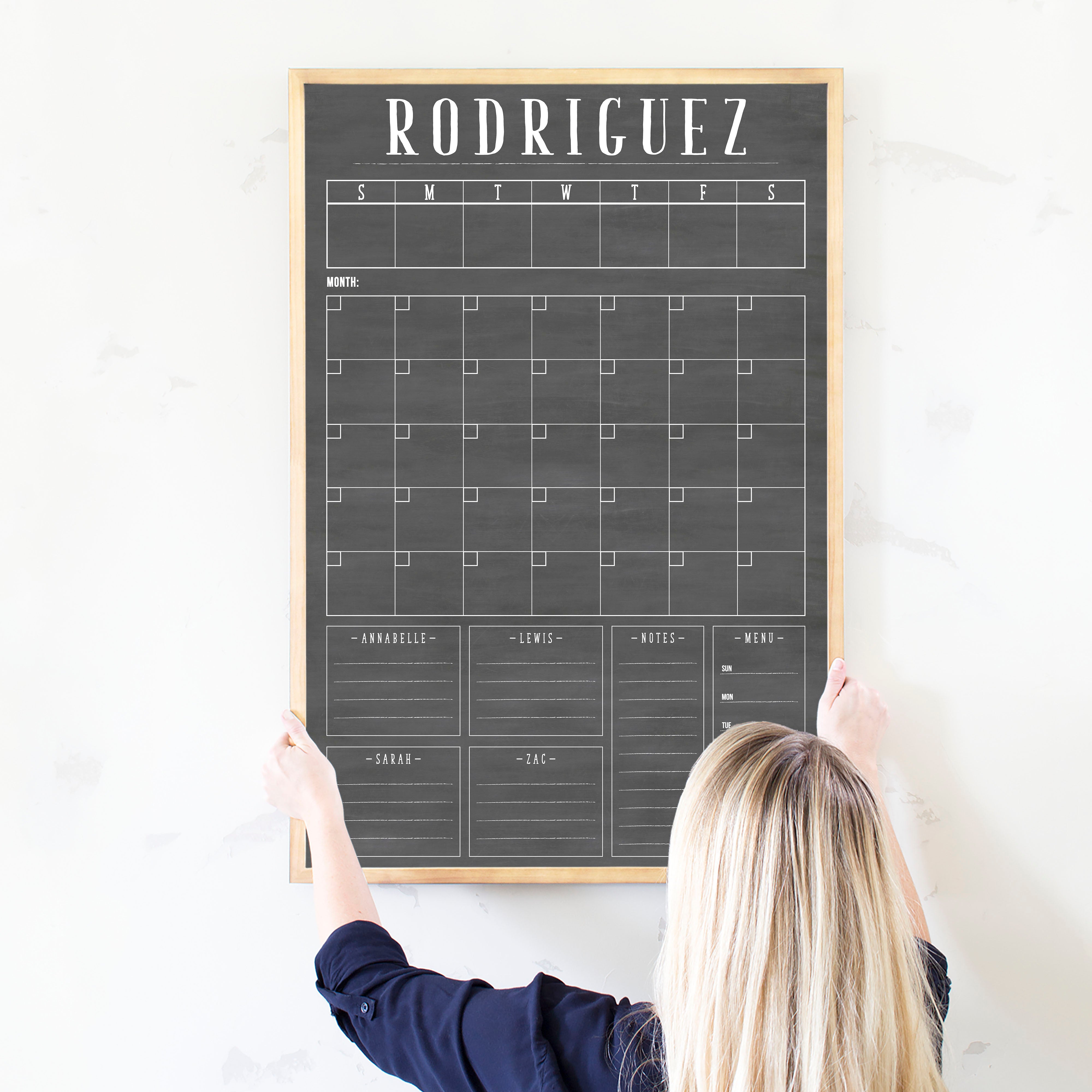 A framed dry-erase chalkboard calendar with a monthly and weekly format hanging on the wall
