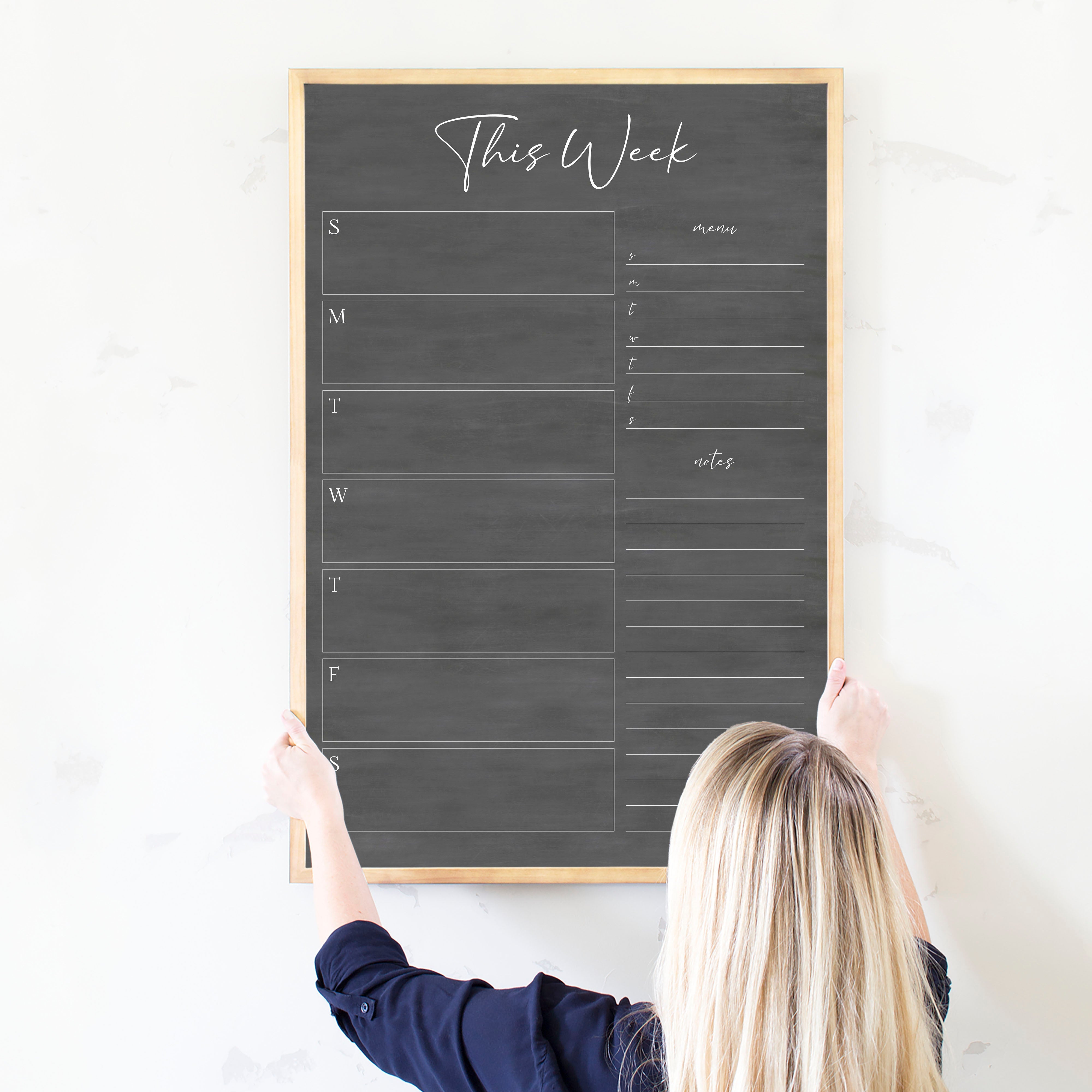 A framed dry-erase weekly calender with a chalkboard look hanging on the wall