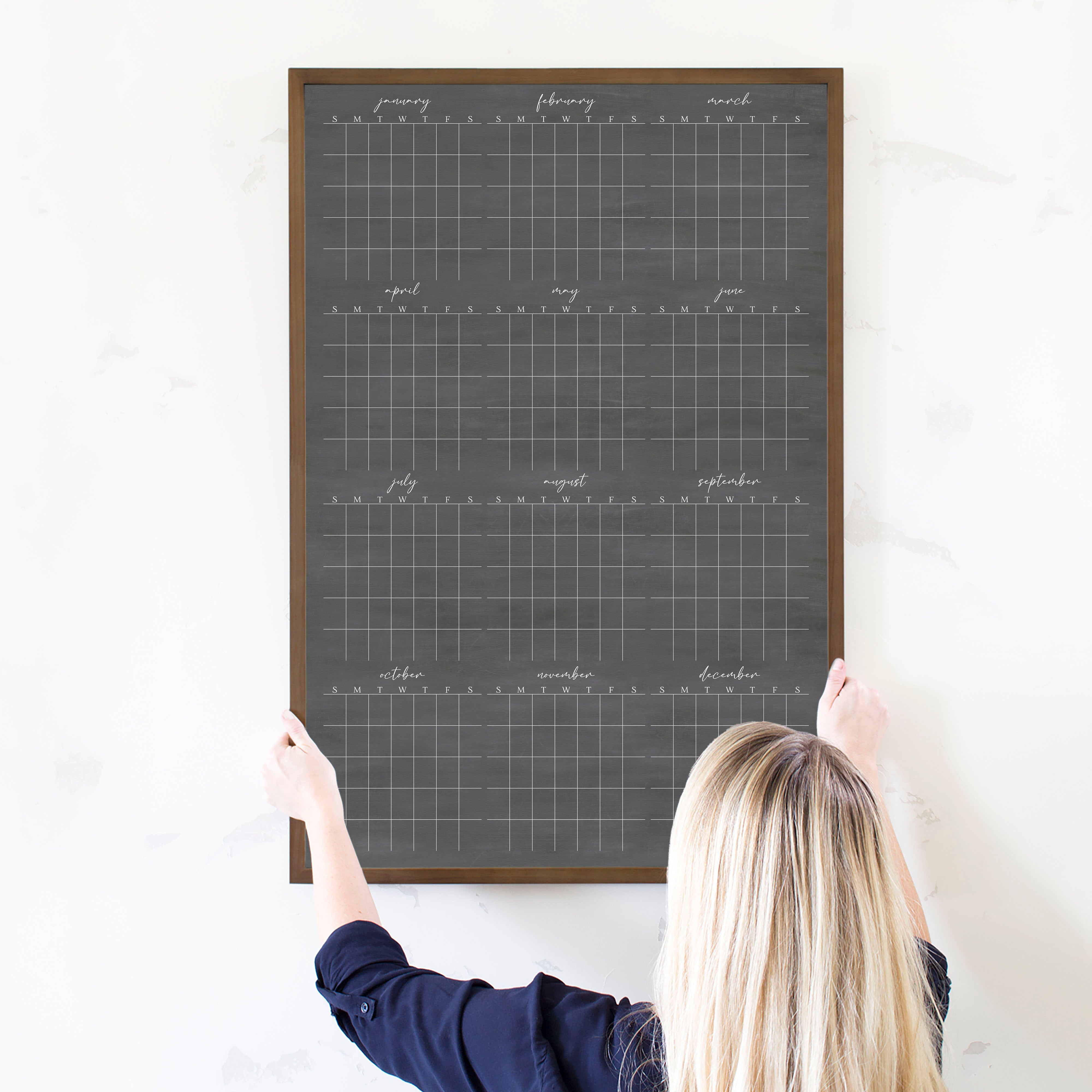 A framed dry-erase yearly calendar with a chalkboard look hanging up on the wall