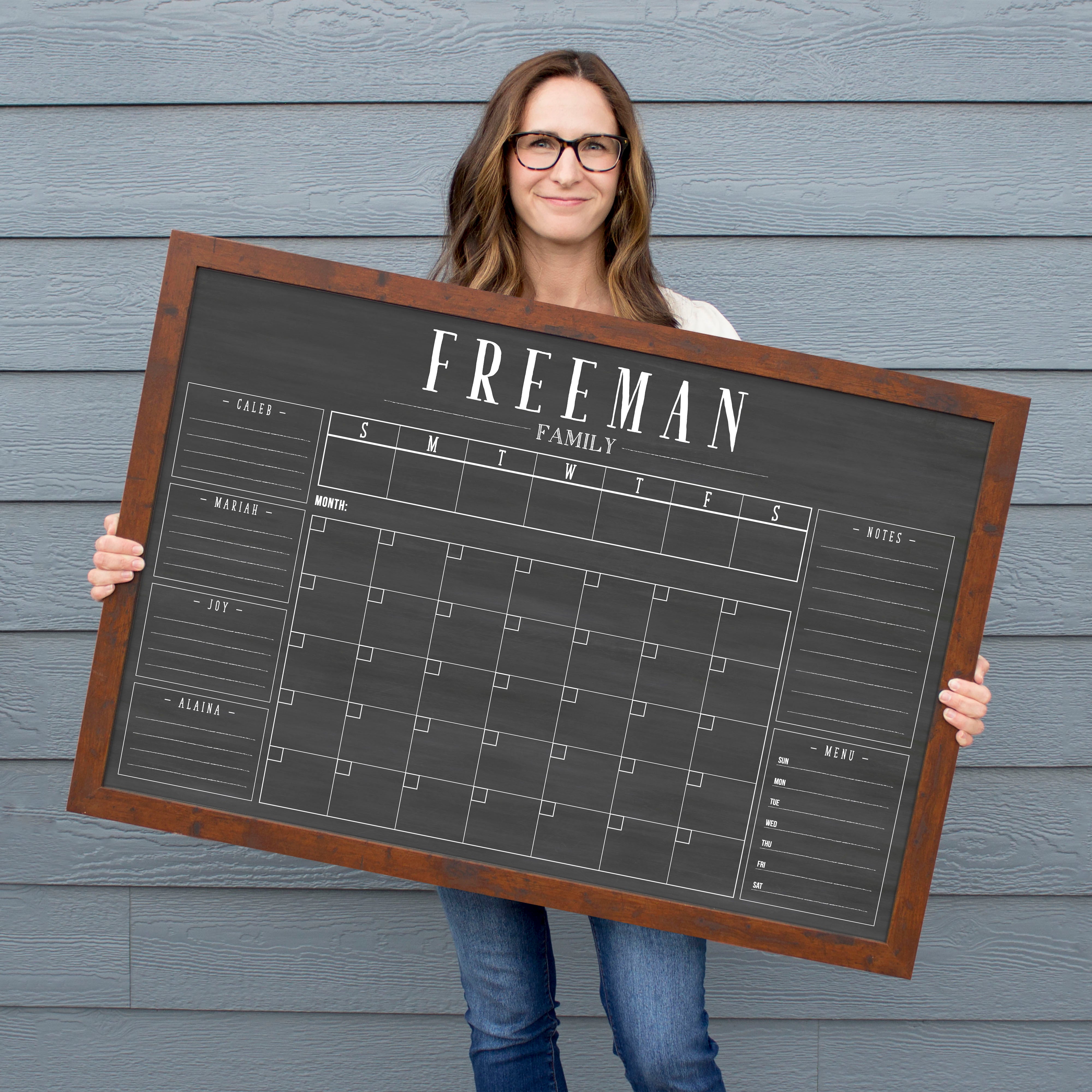A framed dry-erase chalkboard calendar with a monthly and weekly format hanging on the wall