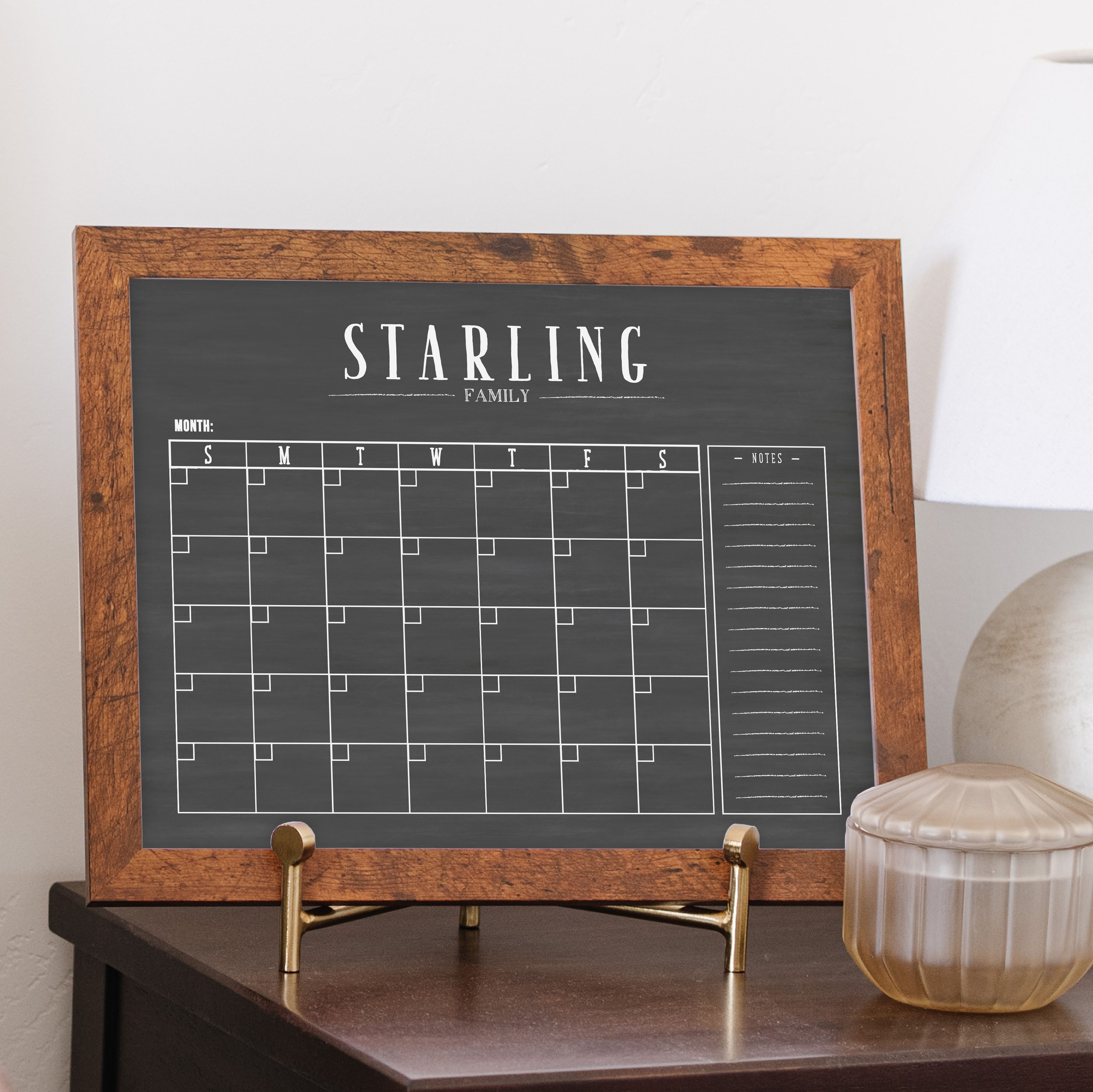 A framed dry-erase monthly calender with a chalkboard look hanging on the wall
